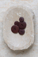 Cinq boutons ronds classiques à deux trous, effet bois veiné brun rouille prune foncé, présentés en tas dans une coupelle en pierre ovale beige marbrée.