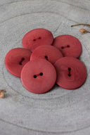 Seven classic two-hole round buttons, terracotta red wood-grain effect, piled on grey stone with a dried leaf in the background.