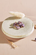 Irregularly shaped, faceted stone-effect buttons in pale mauve purple, with a gold metal shank, piled in a small white ceramic dish on a pink background, with a sprig of dried lagurus grass in the background.