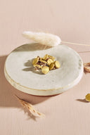 Boutons de forme irrégulière façon pierre facettée, coloris jaune citron doré métallisé, avec fixation à queue métallique dorée, présentés en tas sur une petite coupelle en céramique blanche, sur fond rose avec des épis de lagurus séchés.