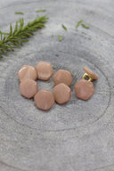 Sept boutons de forme irrégulière façon pierre facettée, aspect nacré, coloris beige rosé sable, avec fixation à queue métallique dorée, posés sur une pierre grise avec un brin de romarin en arrière-plan.