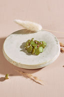 Boutons de forme irrégulière façon pierre facettée, coloris vert matcha olive, avec fixation à queue métallique dorée, présentés en tas sur une petite coupelle en céramique blanche, sur fond rose avec des épis de lagurus séchés.