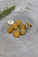 Sept boutons de forme irrégulière façon pierre facettée, aspect nacré, coloris jaune ocre moutarde, avec fixation à queue métallique dorée, posés sur une pierre grise avec un brin de romarin en arrière-plan.