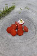 Boutons de forme irrégulière façon pierre facettée, coloris orange vif tangerine, avec fixation à queue métallique dorée, posés sur une pierre grise avec un brin de romarin en arrière-plan.
