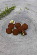 Sept boutons de forme irrégulière façon pierre facettée, coloris brun rouille foncé, avec fixation à queue métallique dorée, posés sur une pierre grise avec un brin de romarin en arrière-plan.