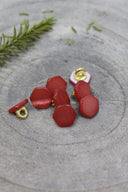 Irregularly shaped, faceted stone-effect buttons in brick-red terracotta, with a gold metal shank, set on grey stone with a sprig of rosemary in the background.