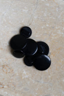 Boutons ronds bombés à queue, sans trous apparents, finition nacrée noir profond brillant, présentés en tas de tailles variées sur une dalle de pierre beige marbrée.