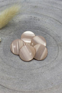 Boutons ronds bombés à queue, sans trous apparents, finition nacrée rose pâle blush, présentés en tas sur une pierre grise, avec un brin d'herbe séchée en arrière-plan.