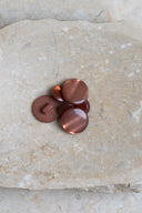 Domed round shank buttons with no visible holes, pearly taupe-pink chestnut finish, piled in various sizes on a slab of marbled beige stone.