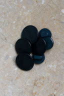 Boutons ronds bombés à queue, sans trous apparents, finition nacrée vert forêt très foncé, presque noir, présentés en tas de tailles variées sur une dalle de pierre beige marbrée.