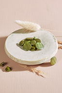 Domed round shank buttons with no visible holes, pearly olive matcha green finish, piled in a small white ceramic dish on a pink background with a dried lagurus spike.