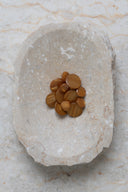 Boutons ronds bombés à queue, sans trous apparents, finition nacrée ocre caramel, présentés en tas de tailles variées dans une coupelle en pierre ovale beige marbrée.