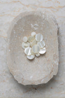 Boutons ronds bombés à queue, sans trous apparents, finition nacrée blanc cassé nacré, présentés en tas de tailles variées dans une coupelle en pierre ovale beige marbrée.