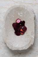 Boutons ronds bombés à queue, sans trous apparents, finition nacrée bordeaux rouille profond, présentés en tas de tailles variées dans une coupelle en pierre ovale beige marbrée.