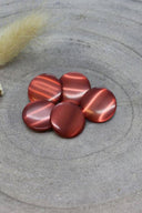 Boutons ronds bombés à queue, sans trous apparents, finition nacrée rouge terracotta, présentés en tas sur une pierre grise, avec un brin d'herbe séchée en arrière-plan.