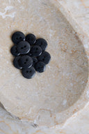 A dozen small glossy two-hole round buttons in deep anthracite black, piled in a marbled beige stone dish.