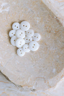 A dozen small glossy two-hole round buttons in pure optic white, piled in a marbled beige stone dish.
