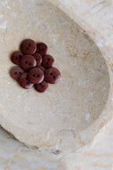 Une douzaine de petits boutons ronds brillants à deux trous, coloris brun rouille bordeaux, présentés en tas dans une coupelle en pierre beige marbrée.