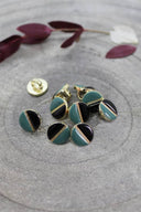 Two-tone round buttons with a gold metal shank, half black and half cactus green, separated by a thin gold trim, piled on grey stone with a sprig of burgundy foliage in the background.