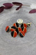 Boutons ronds bicolores à queue métallique dorée, moitié noire et moitié orange rouge tangerine, séparés par un fin liseré doré, présentés en tas sur une pierre grise avec un rameau de feuillage bordeaux en arrière-plan.