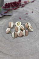 Boutons ronds bicolores à queue métallique dorée, moitié blanc cassé et moitié beige rosé sable, séparés par un fin liseré doré, présentés en tas sur une pierre grise avec un rameau de fleurs séchées bordeaux en arrière-plan.