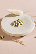 Boutons ronds bicolores à queue métallique dorée, moitié blanc cassé et moitié vert matcha, séparés par un fin liseré doré, présentés en tas sur une petite coupelle en céramique blanche, sur fond rose avec un épi de lagurus séché.
