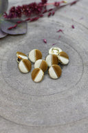 Boutons ronds bicolores à queue métallique dorée, moitié blanc cassé et moitié ocre caramel, séparés par un fin liseré doré, présentés en tas sur une pierre grise avec un rameau de fleurs séchées bordeaux en arrière-plan.