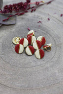 Two-tone round buttons with a gold metal shank, half off-white and half terracotta red, separated by a thin gold trim, piled on grey stone with a sprig of burgundy dried flowers in the background.