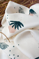 Tissu Palmetto ivoire à motif de feuilles de palmier stylisées marine et rose poudré, plié sur un support en bois devant un fond terracotta.