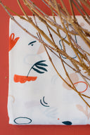 Sandstorm fabric in off-white with an abstract-shape pattern in orange and navy, folded in front of a terracotta background with dried twigs.