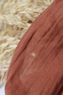 Tissu Sunset terracotta en gaze de coton semi-transparente à motifs soleil brodés fil doré, posé près d'une feuille de palmier.