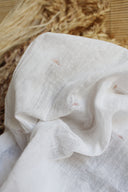 Sunset fabric in off-white, a semi-sheer cotton gauze embroidered with small terracotta sun motifs, placed on a natural-fiber rug near dried wheat stalks.