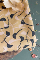 Windy fabric in mustard with a graphic curved-leaf pattern in navy and off-white, folded on a wooden stool with a dried flower and grey, caramel and pink buttons scattered around.