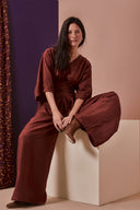 Combi Addie pattern – Woman seated, burgundy jumpsuit with kimono sleeves, tied belt, purple background – Atelier Brunette
