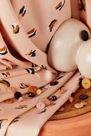 Tissu Beryl Blush drapé autour d'objets en céramique de forme arrondie, avec des boutons roses, terracotta et marine éparpillés sur un plateau en bois.