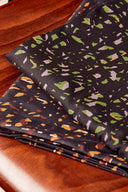 Close-up of the charcoal terrazzo-style confetti-print viscose fabric, with green, lavender, orange and off-white flecks, folded on a wooden table.