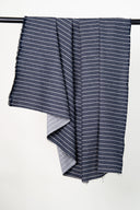 Blue denim fabric with wide ecru stripes, hung on a rail, showing the fabric's light herringbone reverse side, with a frayed selvedge visible.