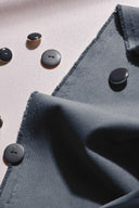 Tissu gabardine légère bleu ardoise plié, lisière effilochée visible, avec des boutons anthracite dispersés autour.