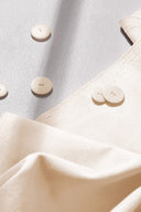 Tissu gabardine légère écru plié, lisière effilochée visible, avec des boutons écru dispersés autour.