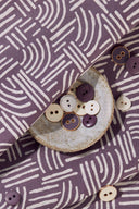 Gros plan sur les plis du tissu Hopscotch mauve à motif de traits entrelacés écru, contre un panier en raphia tressé.