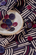 Tissu Hopscotch marine plié, avec une coupelle en céramique contenant des boutons bordeaux, marine et anthracite assortis.