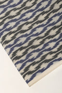 Gros plan sur l'angle plié du tissu écru, détaillant les chevrons bleu et anthracite façon ikat et la lisière écrue.