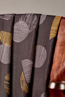 Tissu viscose Jane brun foncé à motif graphique de demi-cercles gris et moutarde, drapé sur l'accoudoir en bois d'une chaise vintage.