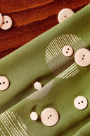 Tissu Jane vert matcha plié avec des boutons écru posés dessus, à côté d'une surface en cuir rouille.