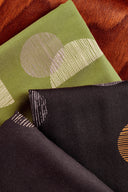 Gros plan sur les tissus viscose Jane pliés, vert matcha et noir, motifs graphiques ronds et rayés, posés sur un fond cuir cognac.