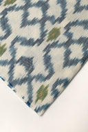 Gros plan sur l'angle plié du tissu écru, détaillant les losanges bleu indigo façon ikat et la lisière effilochée.