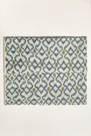 Tissu L'Ikat écru plié à plat, treillis de losanges bleu indigo rehaussé de touches vert olive et orange visible sur l'ensemble de la pièce.