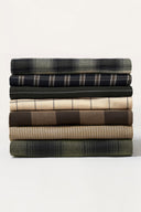 Stack of fabric remnants from the Le Bazaar collection, layered check and stripe patterns in khaki, navy, beige, dark green and brown tones.