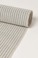 Close-up of the corner of the La Rayure 005 mottled ecru fabric with fine dotted charcoal horizontal stripes, highlighting the pattern and the frayed selvedge.