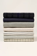 Stack of fabric remnants from the Le Bazaar collection, layered check and stripe patterns in khaki, navy, beige, dark green and brown tones.
